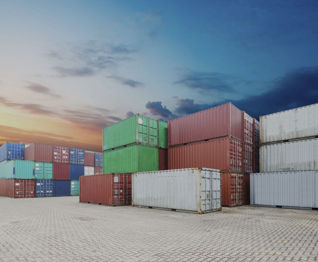 Stacks of colorful shipping containers are arranged on a paved lot under a blue sky with scattered clouds at sunset.