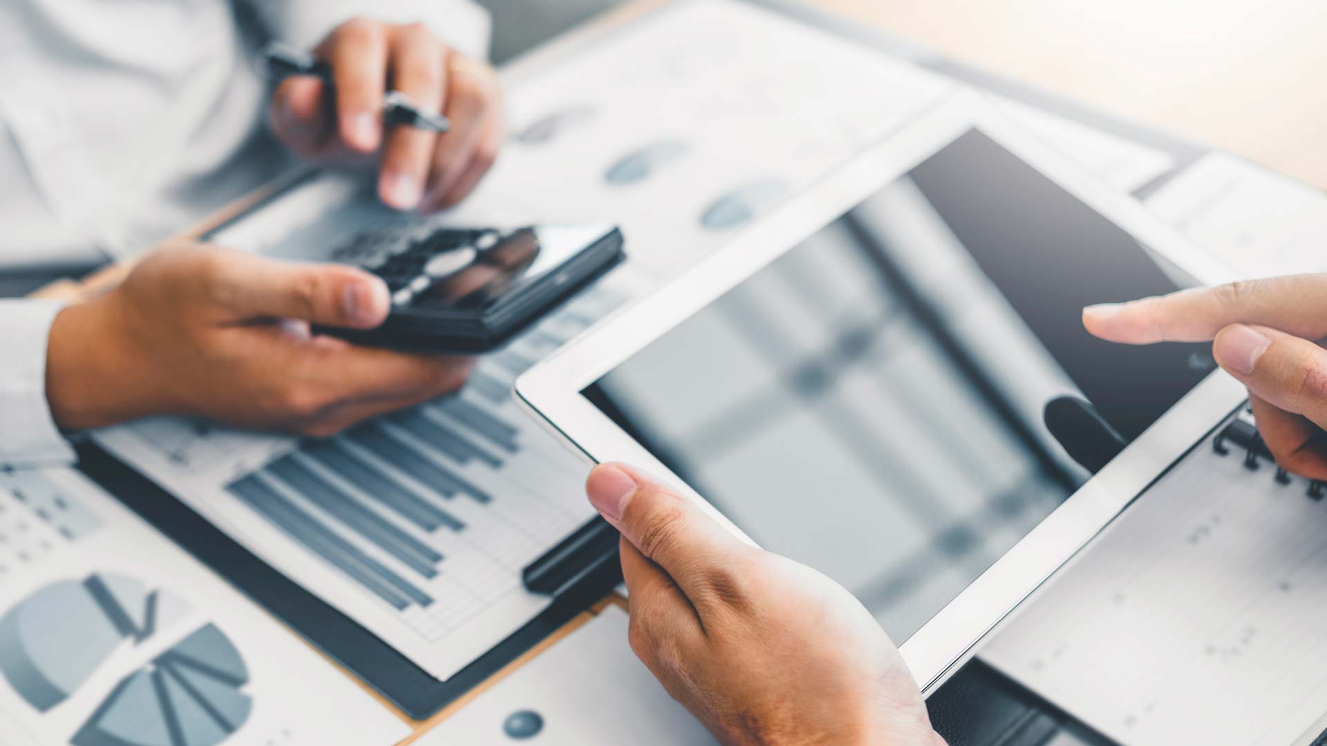 Two people review financial charts on paper; one uses a calculator and pen, while the other holds a tablet with a blank screen and points at it, suggesting a business meeting or financial analysis discussion.