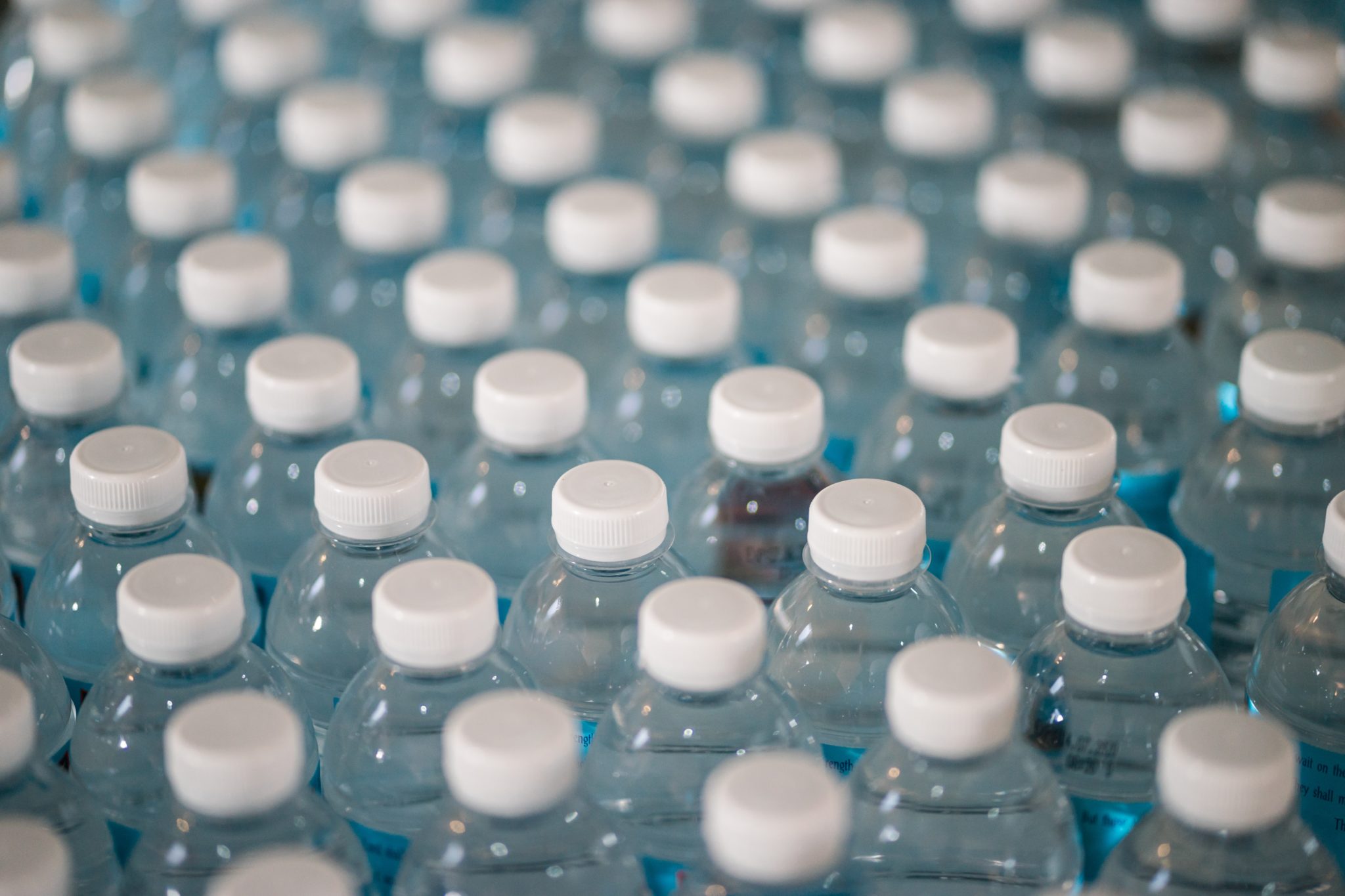 Rows of clear plastic water bottles with white caps are closely packed together, creating a repetitive pattern—ideal for a water delivery service ensuring fresh hydration. The bottles contain clear liquid and are viewed from above.