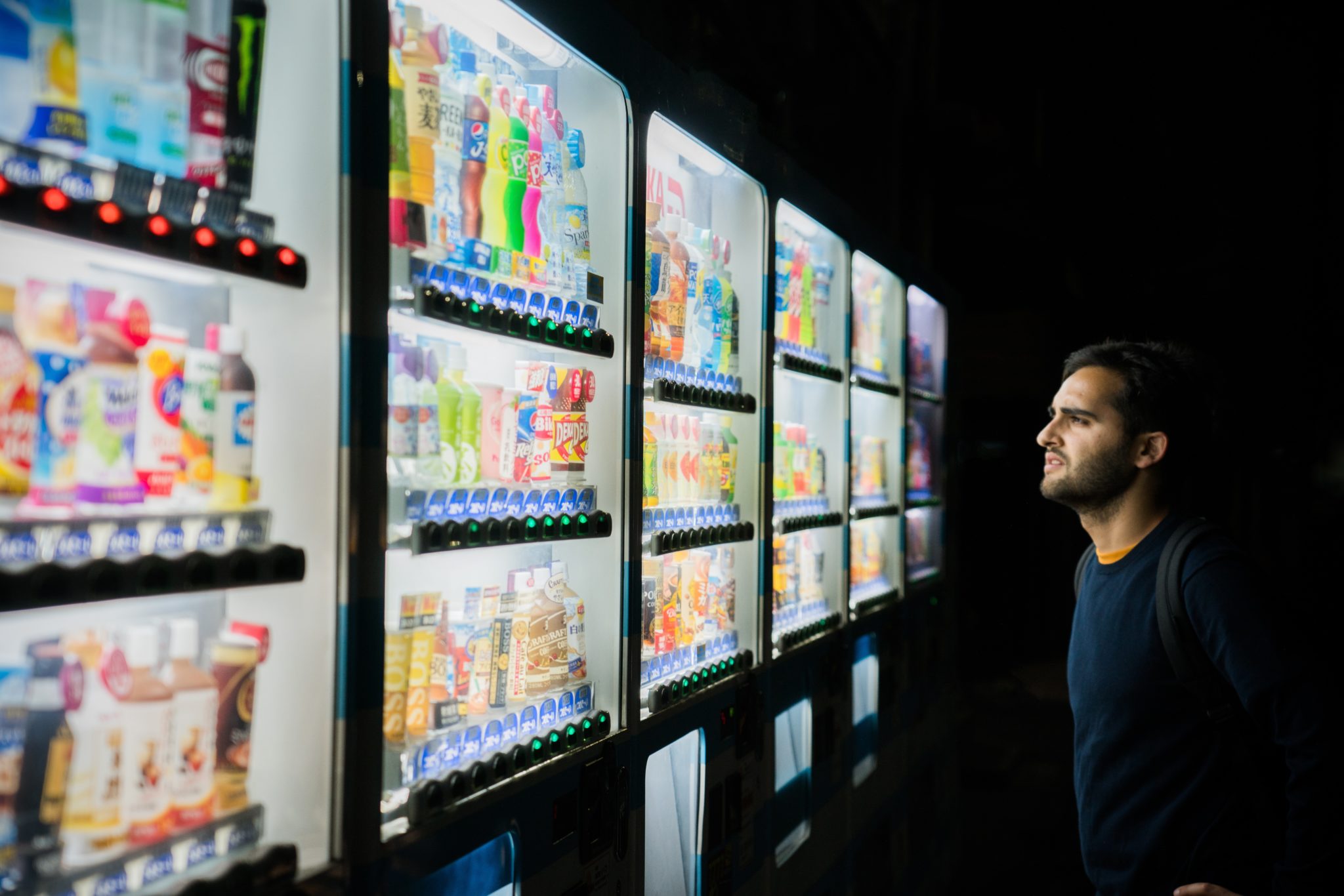 A man stands at night, looking thoughtfully at brightly lit vending machines filled with various drinks and snacks, embodying the paradox of choice.