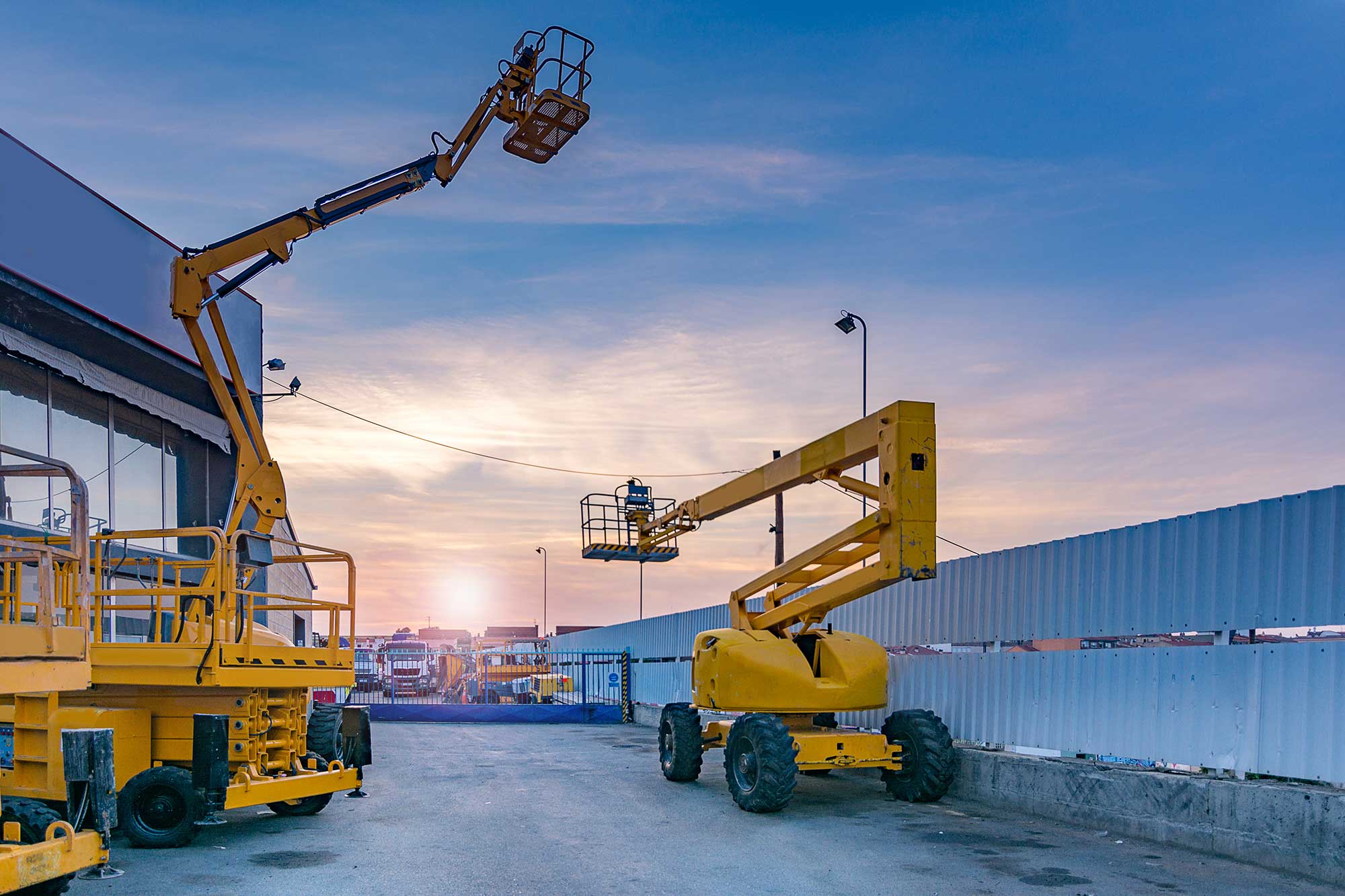 Two yellow boom lifts are parked on a concrete surface near a building at sunset, with one lift extended and a worker standing in its basket against a colorful sky.