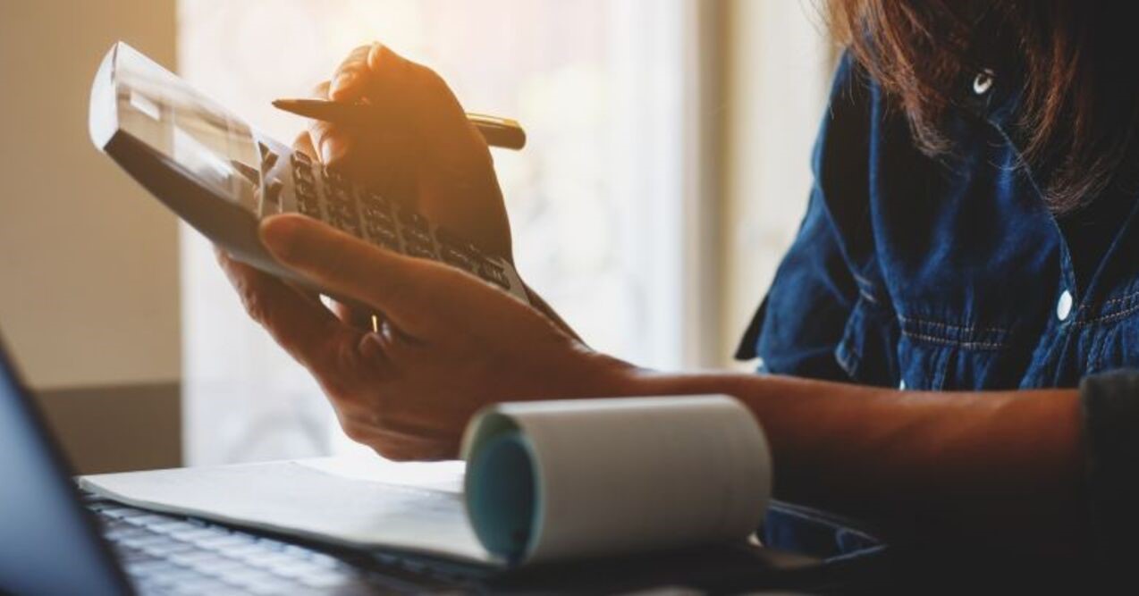 A person in a dark shirt uses a calculator and pen, with a paper roll and laptop nearby, suggesting they are working on finances or using payroll software for accountants. Sunlight streams in from the background.