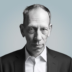 A middle-aged man with short, thinning hair wears a dark suit jacket over a white shirt, looking directly at the camera with a serious expression against a light gray background.