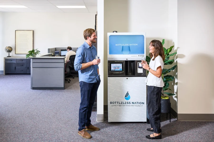 A man and a woman stand and smile while holding cups near a water dispenser labeled Bottleless Nation in a modern office setting. Open workstations and office furniture are visible in the background.