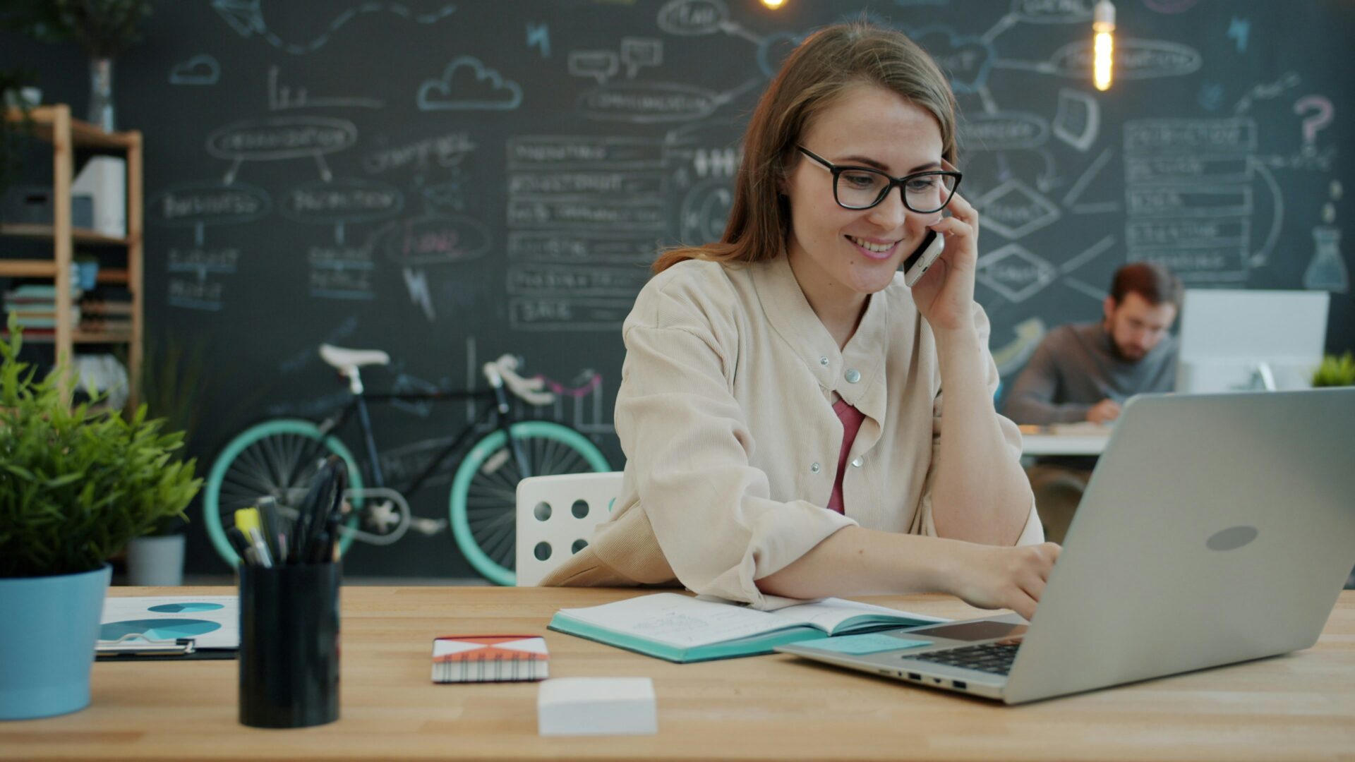 A woman wearing glasses sits at a desk, smiling while talking on the phone and using a laptop. Behind her is a chalkboard wall with drawings and a bicycle. Another person works in the background.