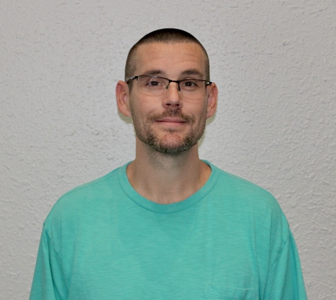 A man with short hair, glasses, and a trimmed beard is standing against a textured white wall, wearing a teal T-shirt and looking at the camera with a neutral expression.
