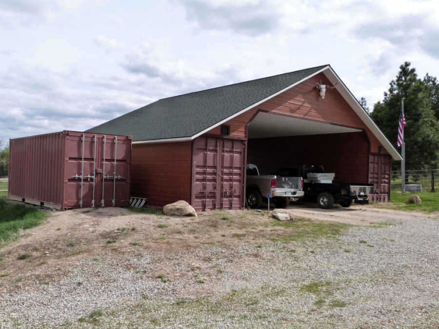 A brown open-sided garage with two pickup trucks parked inside and two large shipping containers placed on either side. An American flag stands to the right, and the scene is outdoors with trees and cloudy skies.
