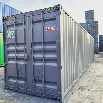 A large gray shipping container with closed doors, labeled with numbers and rental information, sits outdoors on gravel with other stacked containers in the background.